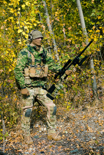 Airsoft man in uniform hold sniper rifle on yellow forest backdrop. Horizontal side view