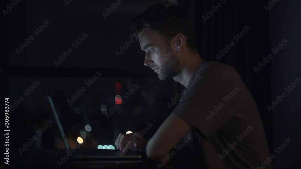 Portrait of Young Man Working Overtime In The Office or Remote Workplace. Using Laptop with Blurred City Light Background in The Window. Late Night Work of Attractive Male. 4K Portrait of Young Man Working Overtime In The Office or Remote Workplace. Using Laptop with Blurred City Light Background in The Window. Late Night Work of Attractive Male. 4K