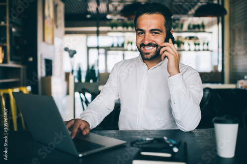 Portrait of successful man in formal wear enjoying smartphone conversation with friend sitting at desktop with modern netbook and smiling at camera, male expert satisfied with distance job indoors