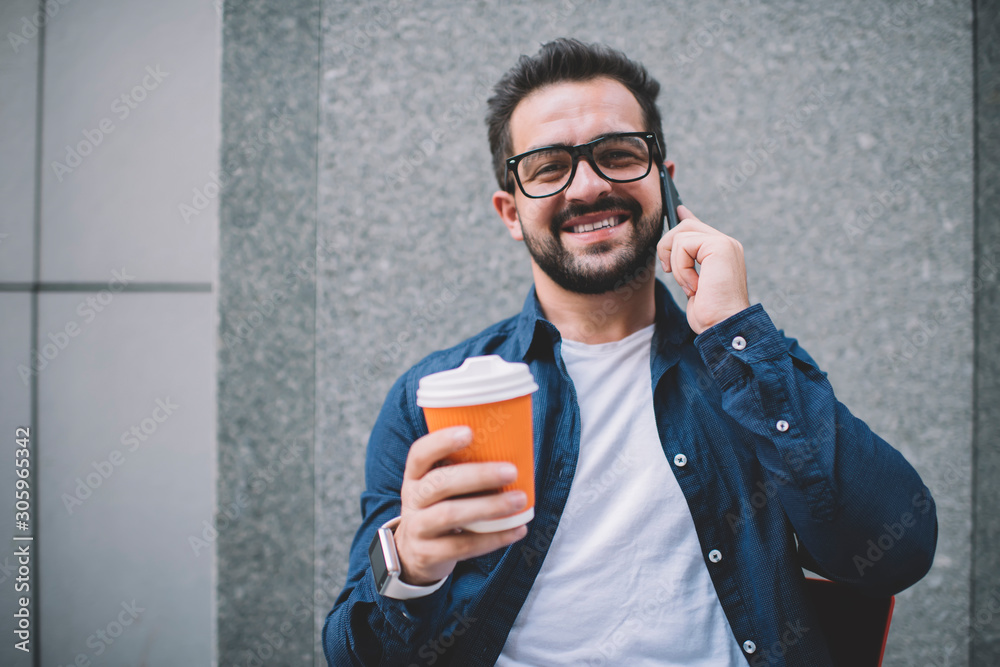 Portrait of cheerful male in classic glasses for vision correction holding takeaway cup with caffeine beverage and making international conversation via cellphone application, roaming connection Portrait of cheerful male in classic glasses for vision correction holding takeaway cup with caffeine beverage and making international conversation via cellphone application, roaming connection