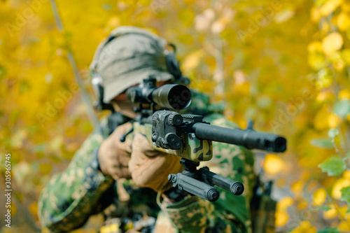 Closeup airsoft man in uniform, stand with sniper rifle on yellow forest backdrop. Soldier aims at the sight