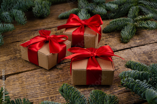 Side view of three handmade boxes with bright red ribbons on a wooden background with vibrant Christmas tree branches.