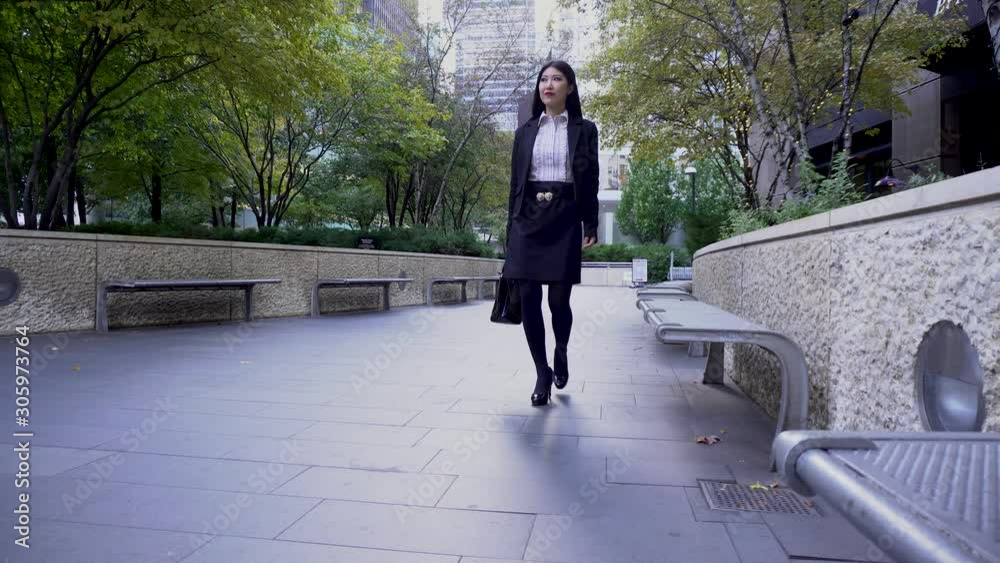 happy Asian business woman with long black hair wearing a trendy black blazer and skirt is walking confidently carrying a fashionable handbag walking along downtown Chicago happy Asian business woman with long black hair wearing a trendy black blazer and skirt is walking confidently carrying a fashionable handbag walking along downtown Chicago