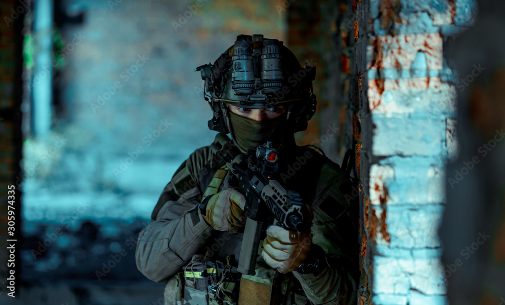Fototapeta premium Man in uniform with machine gun and night-vision device stand beside broken wall. Closeup airsoft soldier in night building. Vertcal photo
