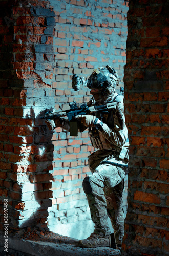 Man in uniform with machine gun and night-vision device move between two broken walls of building. Airsoft soldier in night building