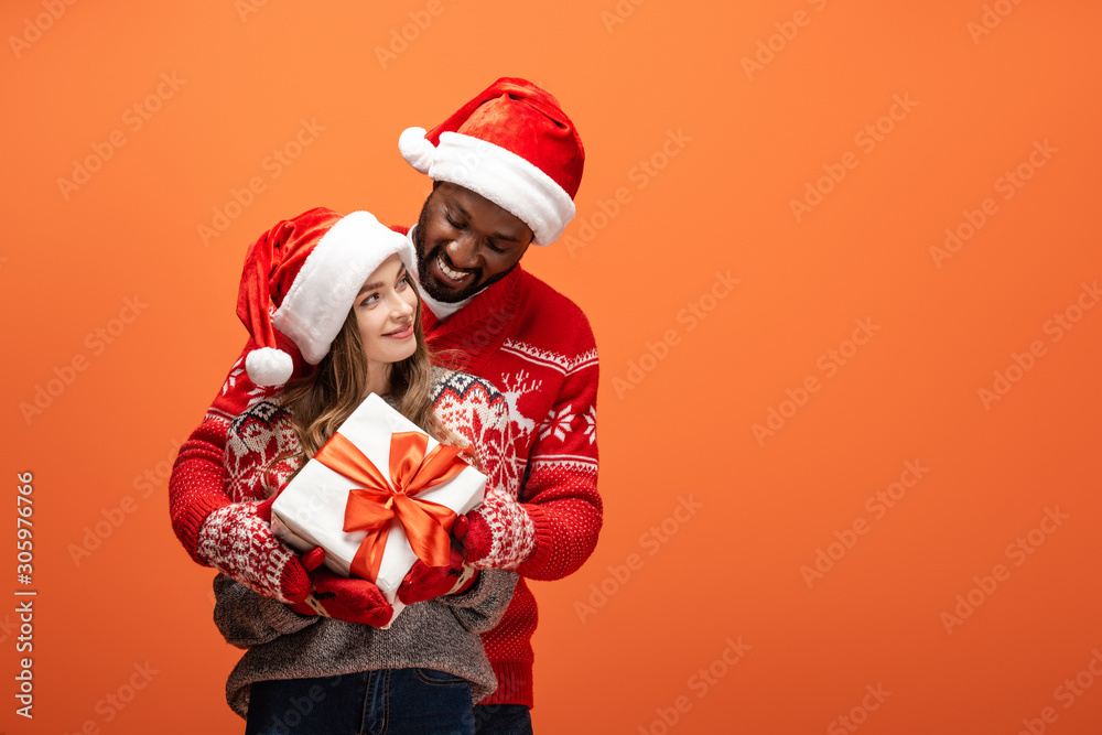 happy interracial couple in santa hats and Christmas sweaters hugging and holding gift on orange background happy interracial couple in santa hats and Christmas sweaters hugging and holding gift on orange background