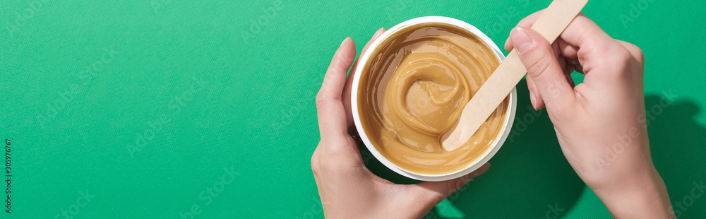 cropped view of woman holding stick and cup with depilation wax on green background, panoramic shot cropped view of woman holding stick and cup with depilation wax on green background, panoramic shot
