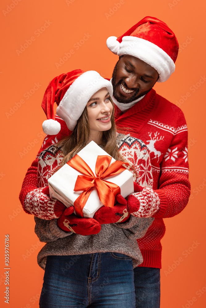 happy interracial couple in santa hats and Christmas sweaters hugging and holding gift on orange background happy interracial couple in santa hats and Christmas sweaters hugging and holding gift on orange background