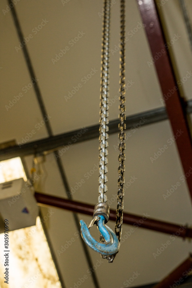 Worn blue hook at the end of a used steel chain connected to the ceiling of a factory used for lifting or winching heavy items across the factory floor saving workers health and safety