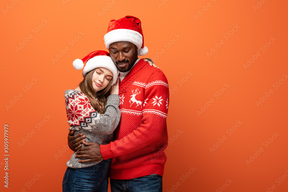 smiling interracial couple in santa hats and Christmas sweaters embracing on orange background