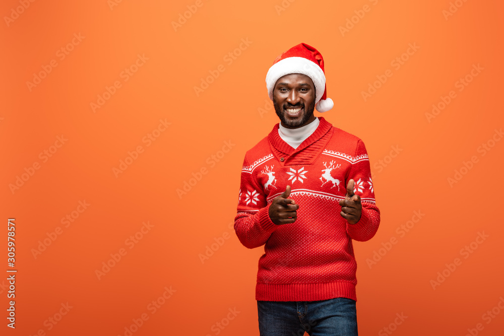 smiling african american man in santa hat and Christmas sweater pointing with fingers on orange background