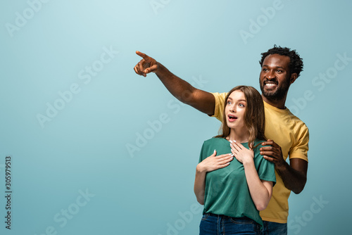 happy and surprised interracial couple pointing with finger away on blue background