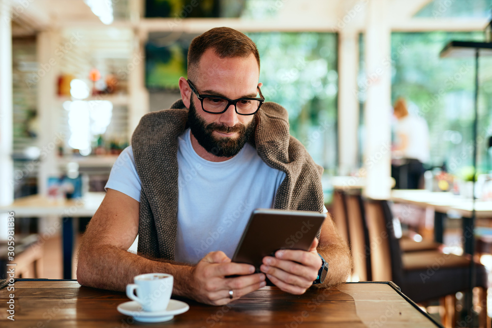 Attractive bearded man with glasses sitting in a cafe searching on tablet Attractive bearded man with glasses sitting in a cafe searching on tablet