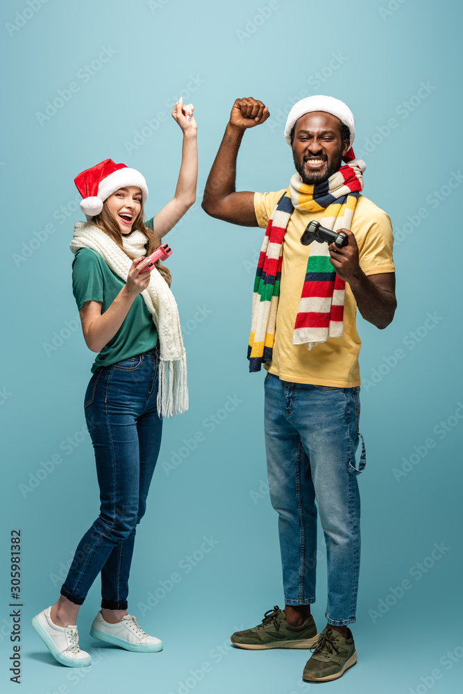 KYIV, UKRAINE - AUGUST 22, 2019: excited interracial couple in santa hats and scarves holding joysticks and showing yes gesture