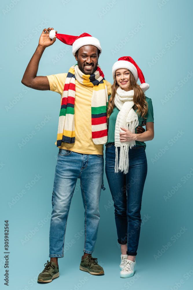 happy interracial couple in santa hats and scarves on blue background happy interracial couple in santa hats and scarves on blue background