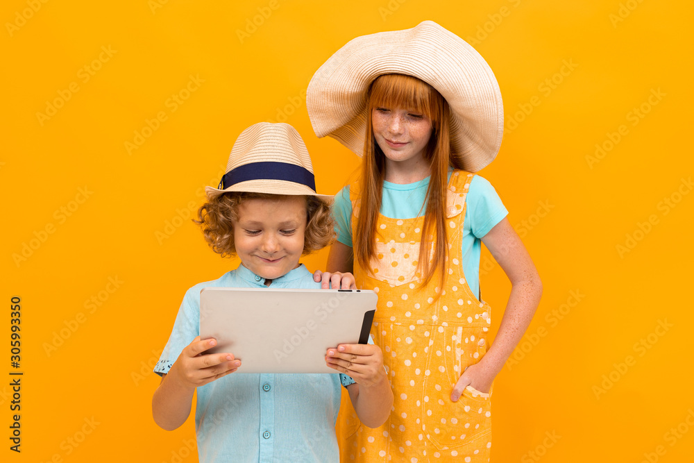 Red-haired brother and sister in summer hats book tickets on a tablet on a yellow background.