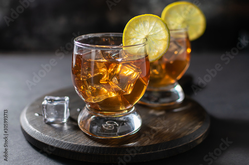 Two glasses of scotch or bourbon with ice and lime on the wooden tray on dark vintage background. Party drinks concept.