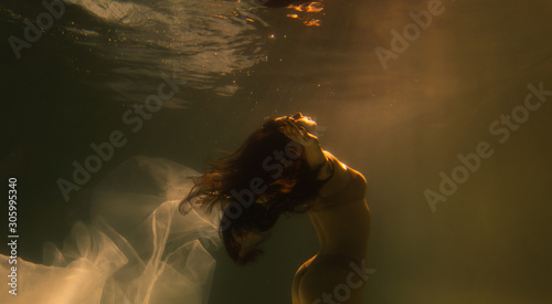 Beautiful girl swims underwater with long hair. Blue or gold background like gold. The atmosphere of a fairy tale or magic. Diving under the water with a shiny cloth