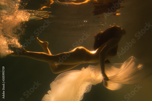 Beautiful girl swims underwater with long hair. Blue or gold background like gold. The atmosphere of a fairy tale or magic. Diving under the water with a shiny cloth