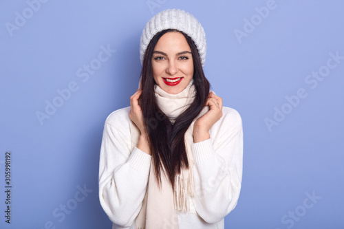 Beautiful brunette woman with long hair wearing stylish warm cap and white sweater, looking directly at camera, keeps hands on her scarf, female with bright make up, standing against blue studio wall.