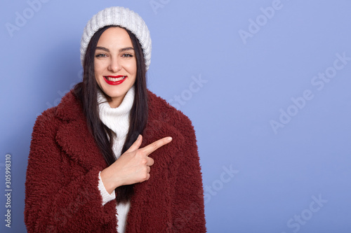 Smilng young brunette woman with long dark hair and red pomade,female dresses faux fur coat and white winter cap pointing aside with her fore finger, looking at camera, standing over blue background. photo