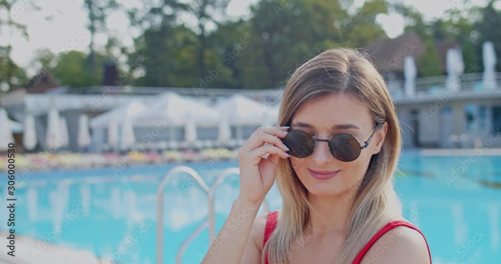 Close up of the pretty Caucasian young woman taking on sunglasses and smiling joyfuly to the camera at the swimming pool. Portrait. Close up of the pretty Caucasian young woman taking on sunglasses and smiling joyfuly to the camera at the swimming pool. Portrait.