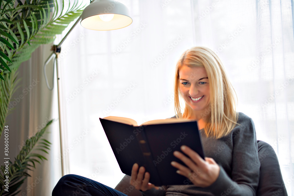 Cheerful blonde woman sitting on her couch reading a book at home in the sitting room Cheerful blonde woman sitting on her couch reading a book at home in the sitting room