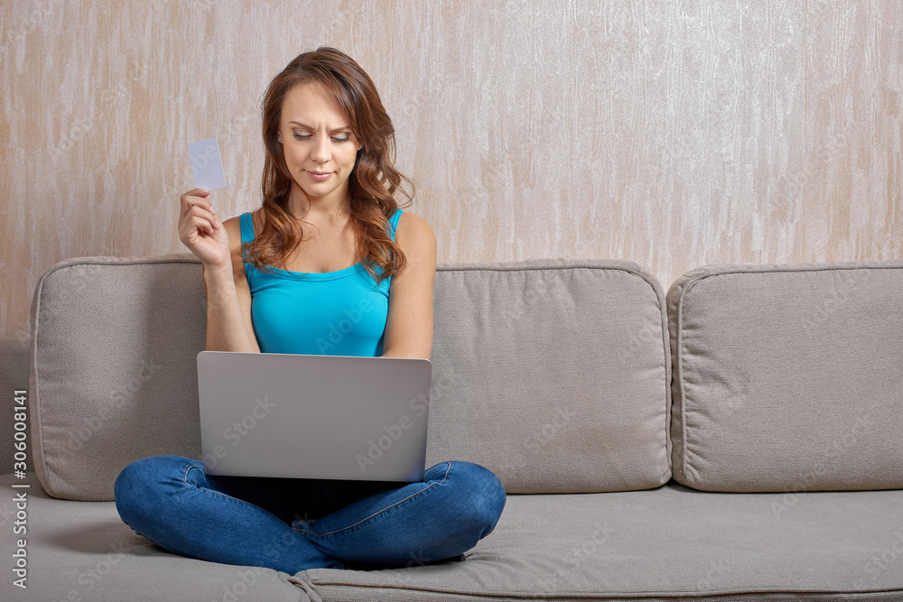 Beautiful woman wearing cyan top with thin shoulder straps and jeans sitting cross-legged on cushions sofa looking at the screen of laptop on her lap. Beautiful woman wearing cyan top with thin shoulder straps and jeans sitting cross-legged on cushions sofa looking at the screen of laptop on her lap.