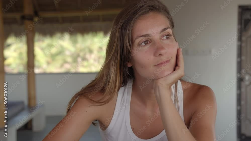 Tilt down shot of young Caucasian woman thinking and typing on laptop while doing freelance work in tropical patio during vacation