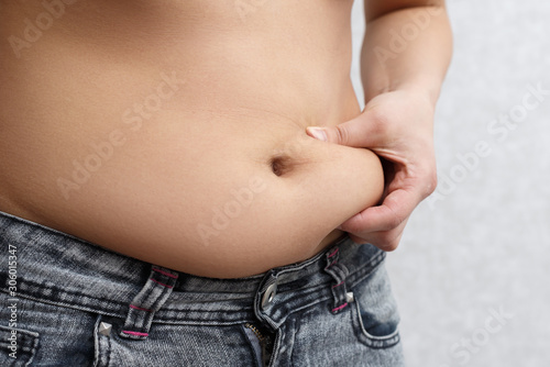 the woman's hand holds a fold of excess fat on her stomach. on grey background, side view