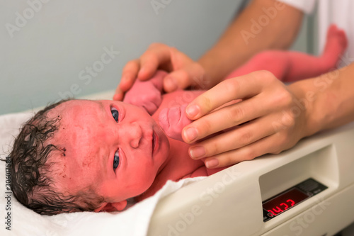 New born baby on a weight, looking directly to the camera - midwife's hand keep care on baby - closeup photo