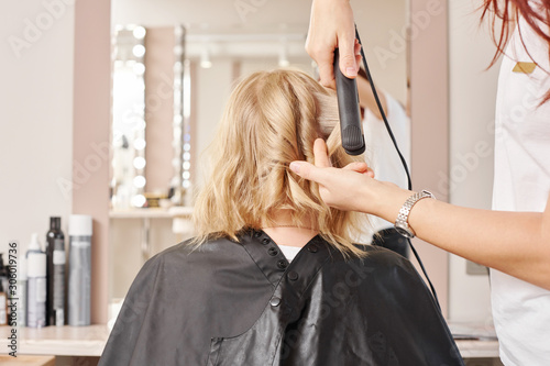 Female hairdresser standing and making hairstyle to blonde woman in beauty salon