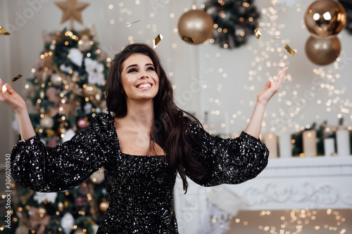 Portrait of a beautiful girl in a black dress at Christmas interior. Smiling young woman in a festive dress with a Christmas gifts and Christmas tree. Happy New Year 2020.
