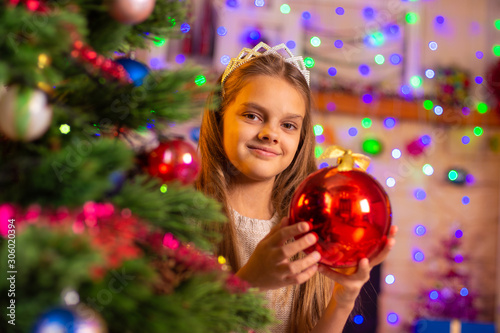 Beautiful ten year old girl peeking out with a big red ball from behind a Christmas tree