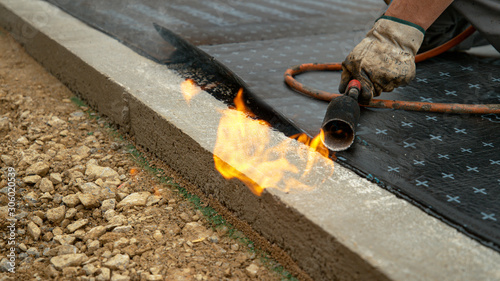 CLOSE UP: Contractor wielding a torch burns a roll of tar over the foundation.