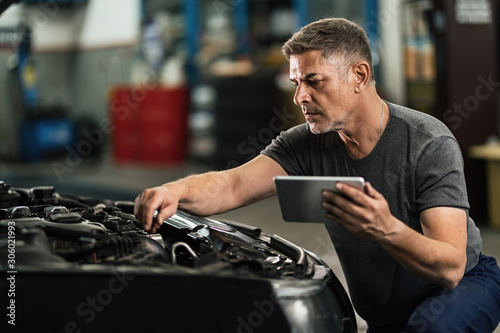 Car mechanic using digital tablet while examining engine in auto repair shop.