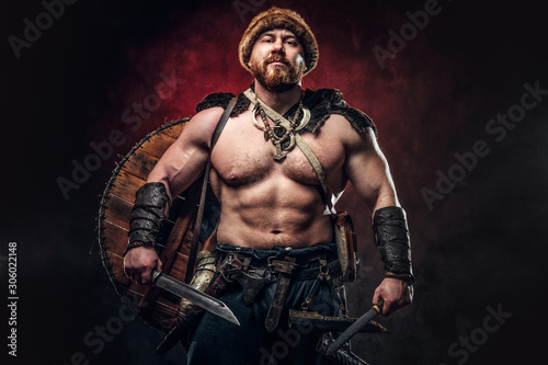 Serious Viking clad in light armor with a shield behind his back holds a sword and an axe. Posing on a dark background with red light