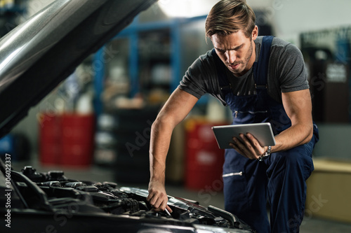 Young auto mechanic using touchpad while repairing car engine in a garage. © Drazen