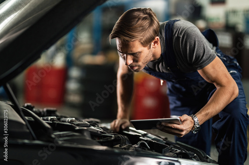 Young mechanic examining broken car engine while using touchpad in auto repair shop.