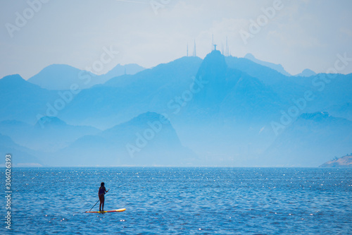 Woman stand up paddle on Camboinhas beach in Niteroi, surfing with Christ the Redeemer statue and the mountains of Rio de Janeiro in the background, clouds separating the mountains from the blue sea.