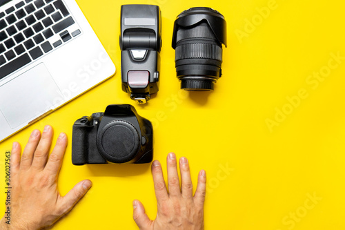 Photo camera and other accessories such as flash light and lens are on the table near laptop on yellow background. Man photographer prepairs photo for editing. View from the top, flat lay. photo