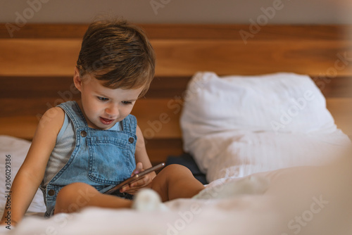 Cute little kid focused on smartphone while sitting on the bed. Boy playing games on smartphone. Social and technology concept. Casual baby watching a mobile phone. Baby boy playing with smartphone