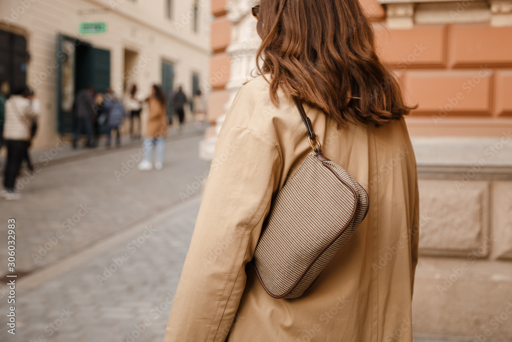street style fashion details. an attractive woman wearing a beige trench  coat and 90s pattern vintage shoulder mini bag, crossing the street.  fashion outfit perfect for autumn Stock Photo