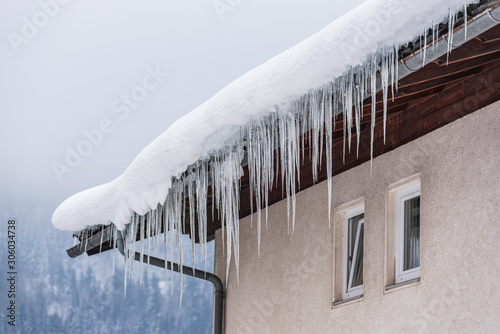 Big icicles and snow hanging over the rain gutter on a roof of a traditional wooden house in the mountains in winter could be dangerous. Blue sky at the background.