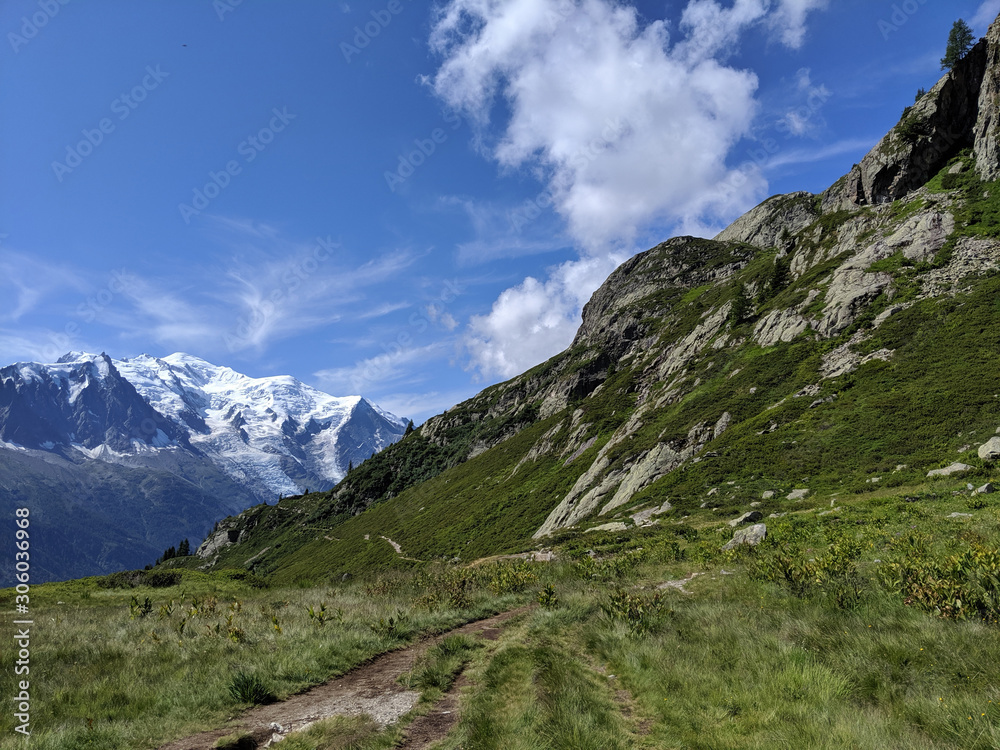 A scenic Alpine view of the famous trek Tour du Mount with sunny blue sky, a few clouds and snowy mountains far away