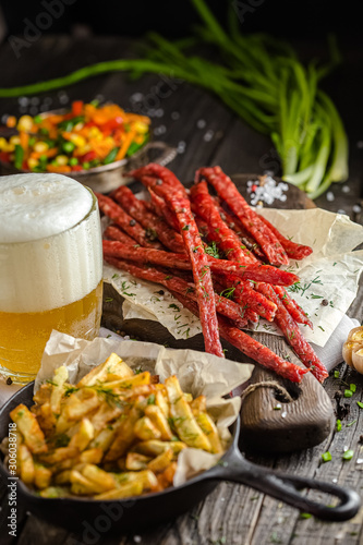 meat appetizer,  snacks, thin sausages dried for beer, lie on a dark wooden and cutting board table, fresh herbs, a glass of beer and foam, fried potatoes in a pan