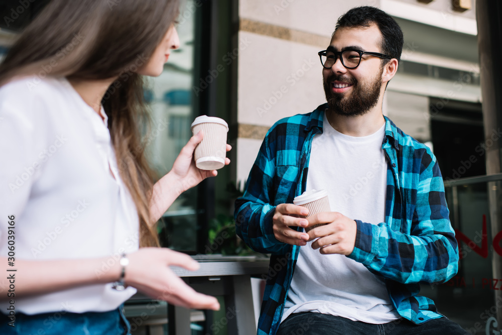 Smiling asian male laughing during conversation with caucasian female colleague during coffee break, happy hipsters friends talking to each other holding takeaway cup and telling funny stories Smiling asian male laughing during conversation with caucasian female colleague during coffee break, happy hipsters friends talking to each other holding takeaway cup and telling funny stories