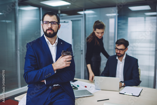 Thoughtful business man pondering in office during coffee break with professional colleagues on background, bearded financial director in optical spectacles for provide eyes protection in office