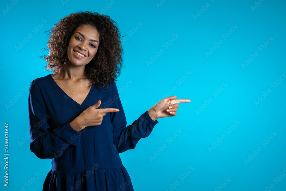 Portrait of girl, she indicates with arms on her left with copy space. Happy smiling african woman presenting and showing something isolated on blue background.