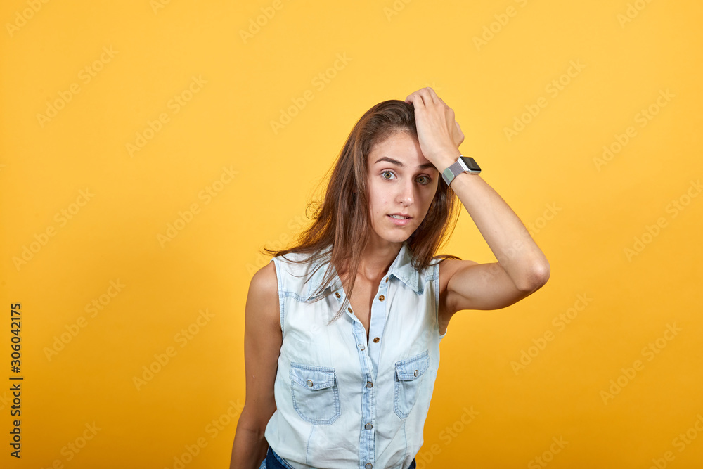 Confused caucasian young woman in blue denim shirt keeping hand on head, looking sad isolated on orange background in studio. People sincere emotions, lifestyle concept. Confused caucasian young woman in blue denim shirt keeping hand on head, looking sad isolated on orange background in studio. People sincere emotions, lifestyle concept.